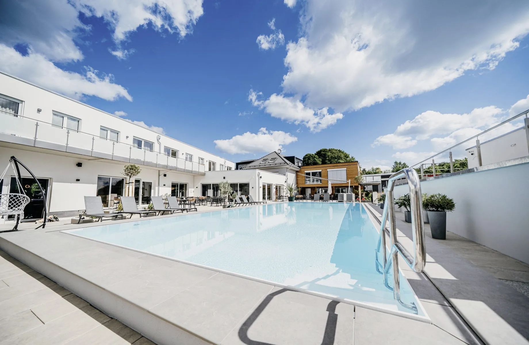 Outdoorpool des Familienhotel Friedrichshof, umgeben von weißem Hotelgebäude, Liegen, Hängesessel, Pflanzen unter blauem Himmel mit Wolken