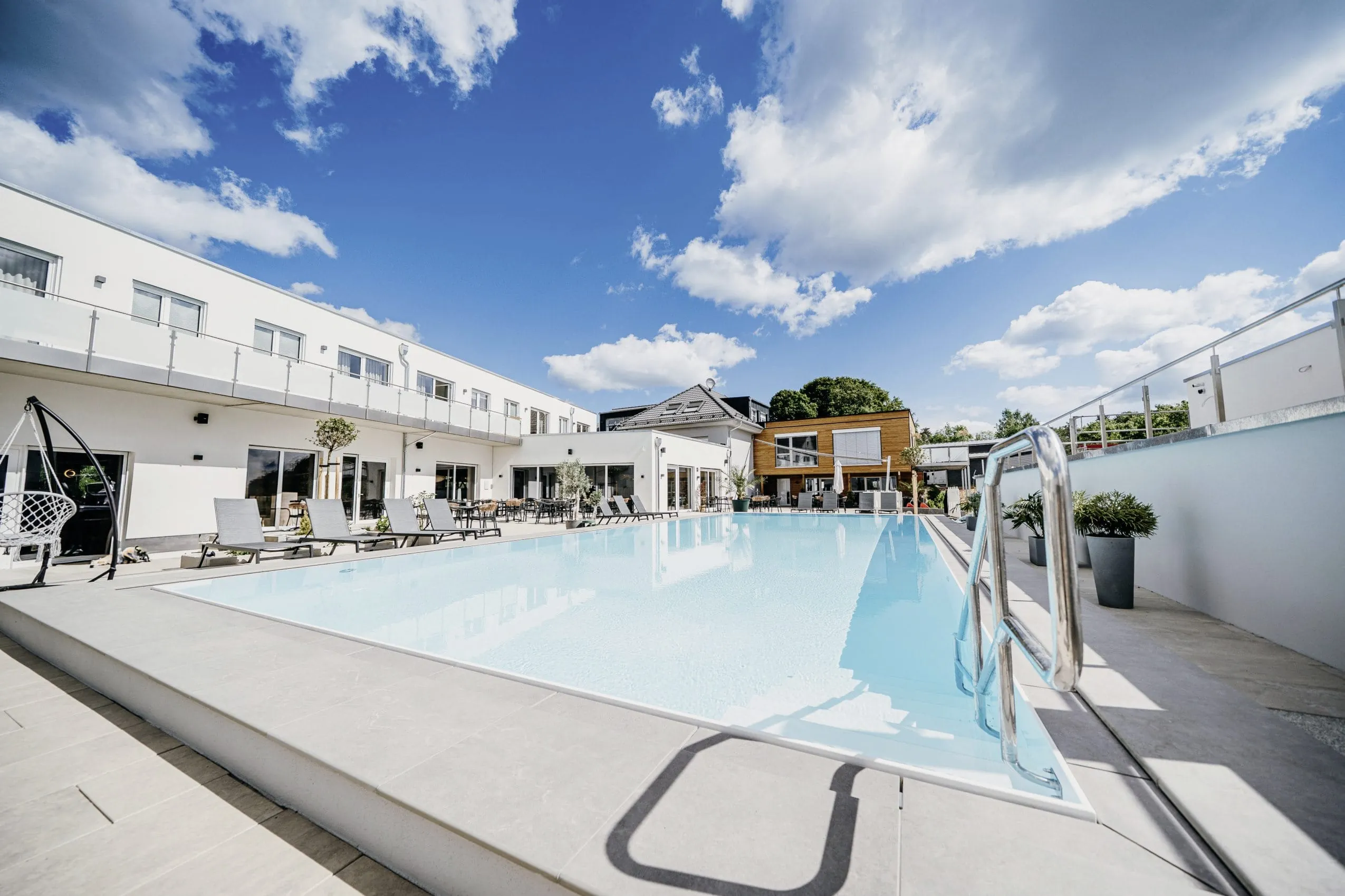 Outdoorpool des Familienhotel Friedrichshof, umgeben von weißem Hotelgebäude, Liegen, Hängesessel, Pflanzen unter blauem Himmel mit Wolken