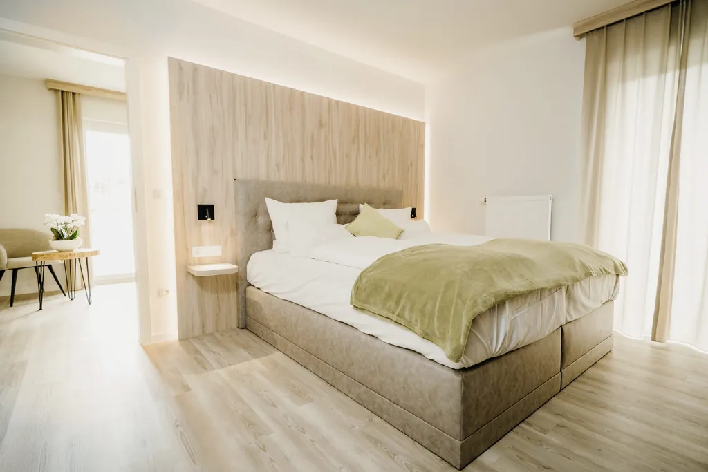 Hell eingerichtetes Schlafzimmer im Familienhotel Friedrichshof mit großem Bett, Holzpaneel hinter dem Kopfteil und Fenster mit Vorhängen