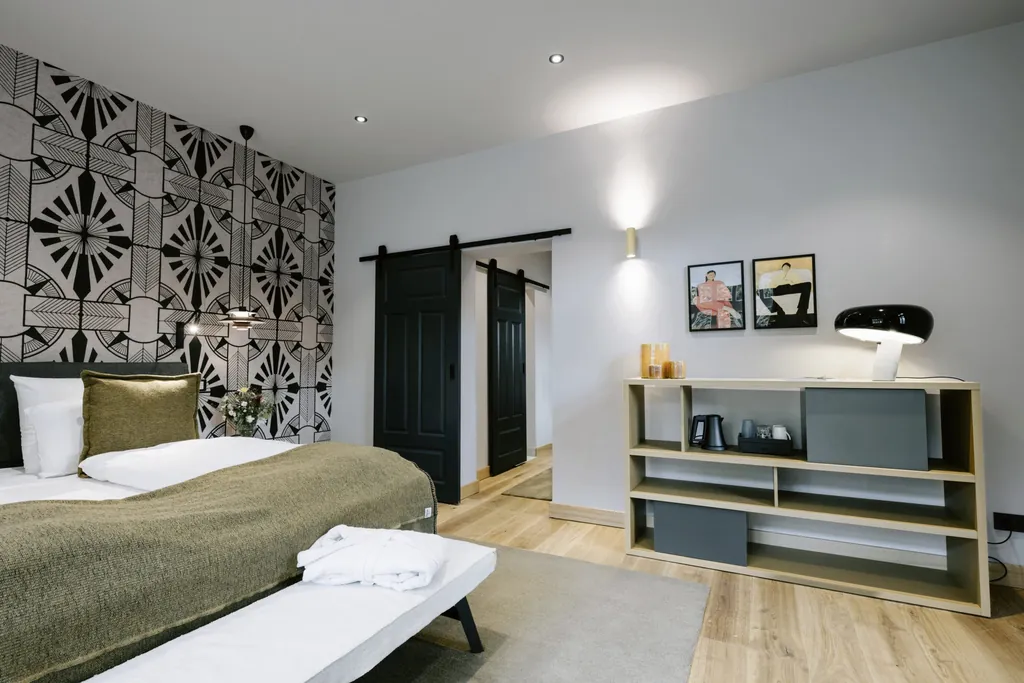 Modern eingerichtetes Schlafzimmer im Familotel Landhaus Averbeck mit gemusterter Wandtapete, schwarzer Schiebetür, Holzregal mit grauen Boxen und schwarzer Tischlampe