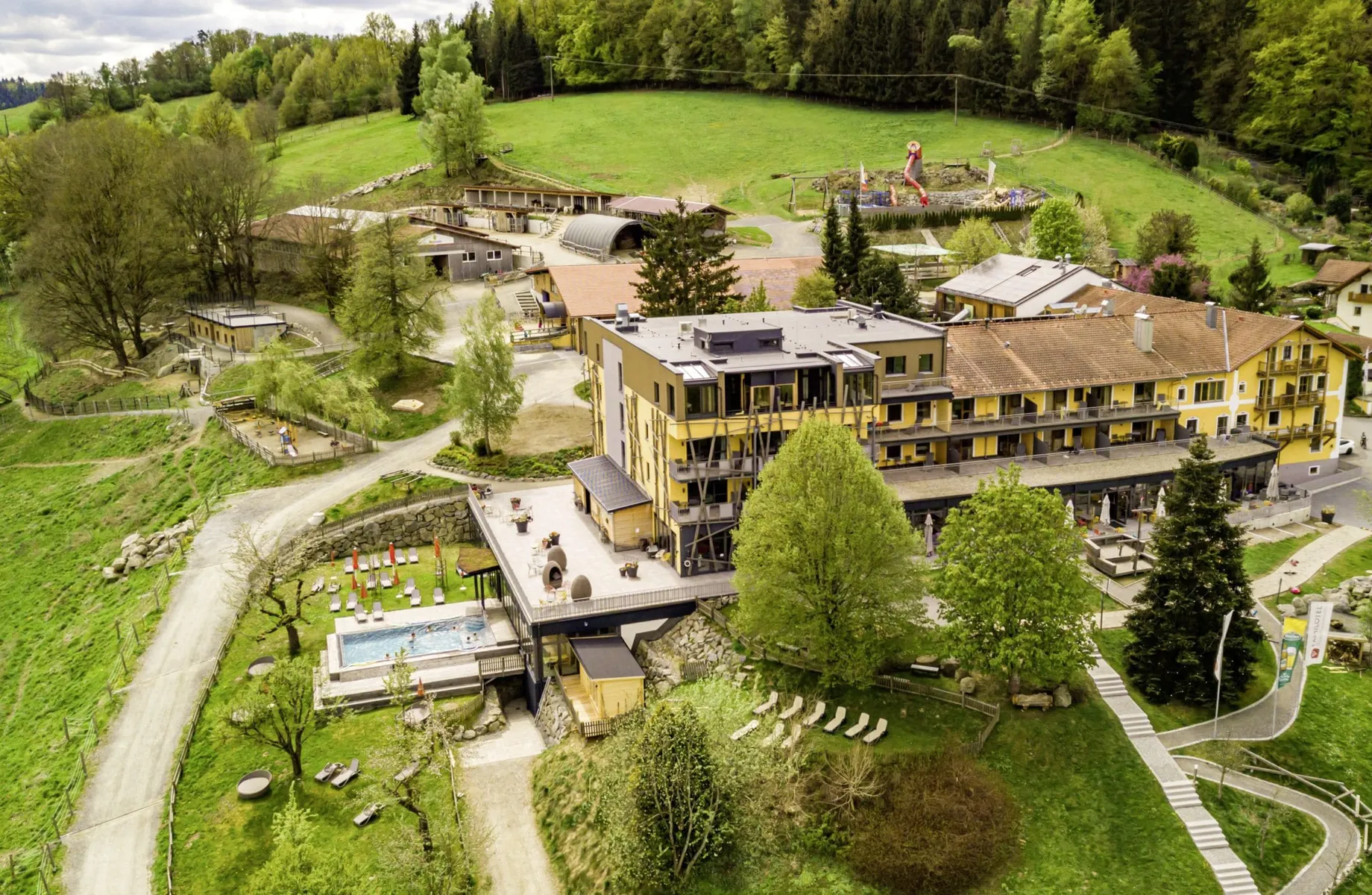 Luftaufnahme des Hotel & Resort Landhaus zur Ohe mit gelb gestrichener Fassade, Reitanlage, Spielplatz, Outdoorpool, Liegen, umgeben von Bäumen und Wiesen