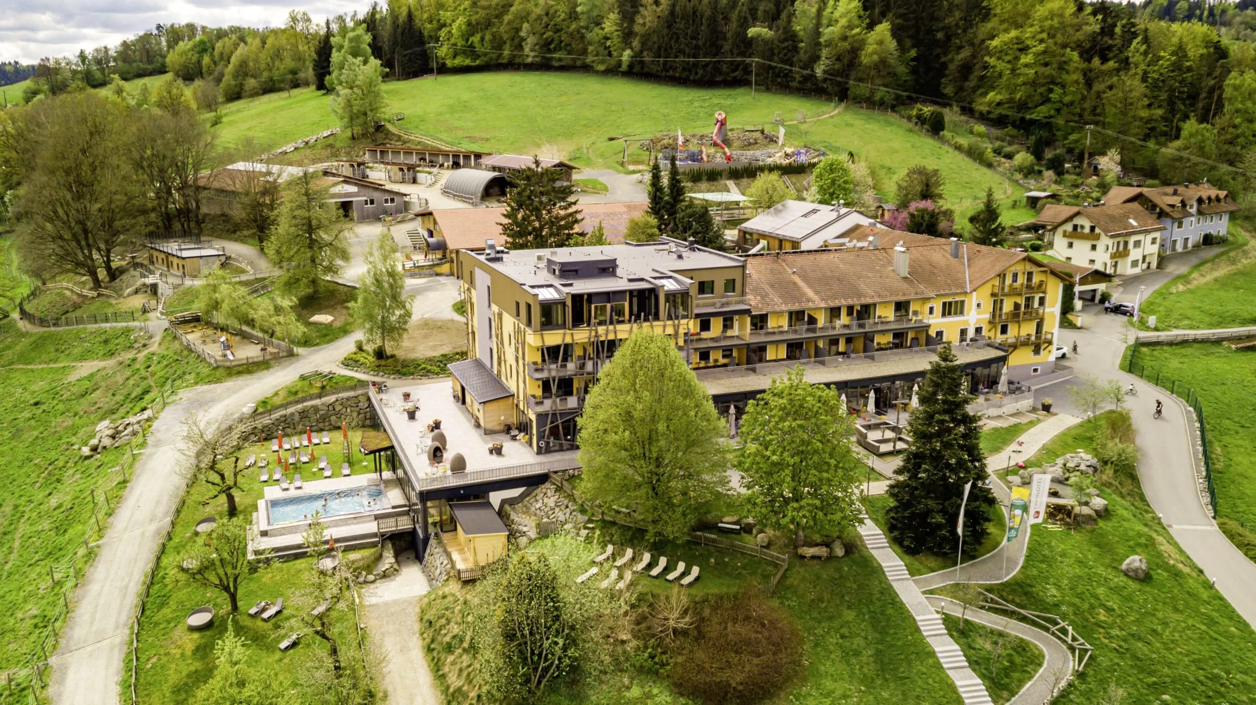 Luftaufnahme des Hotel & Resort Landhaus zur Ohe mit gelb gestrichener Fassade, Reitanlage, Spielplatz, Outdoorpool, Liegen, umgeben von Bäumen und Wiesen
