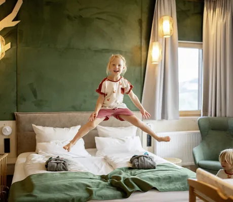 Blondes Mädchen springt auf einem Doppelbett mit grünem Überwurf in einem Zimmer des Hotel & Resort Landhaus zur Ohe mit grüner Wand und grünem Sessel
