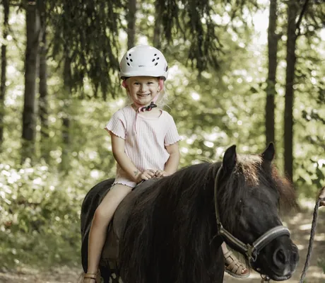 Mädchen mit weißem Helm und rosa gestreiftem T-Shirt sitzt auf einem schwarzen Pony im Wald in der Nähe des Familotel Schreinerhof