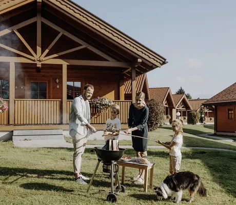 Familie mit Hund steht vor einem Chalet des Chaletdorfs ZellerSee auf Rasen und grillt Würstchen an einem Holzkohlegrill