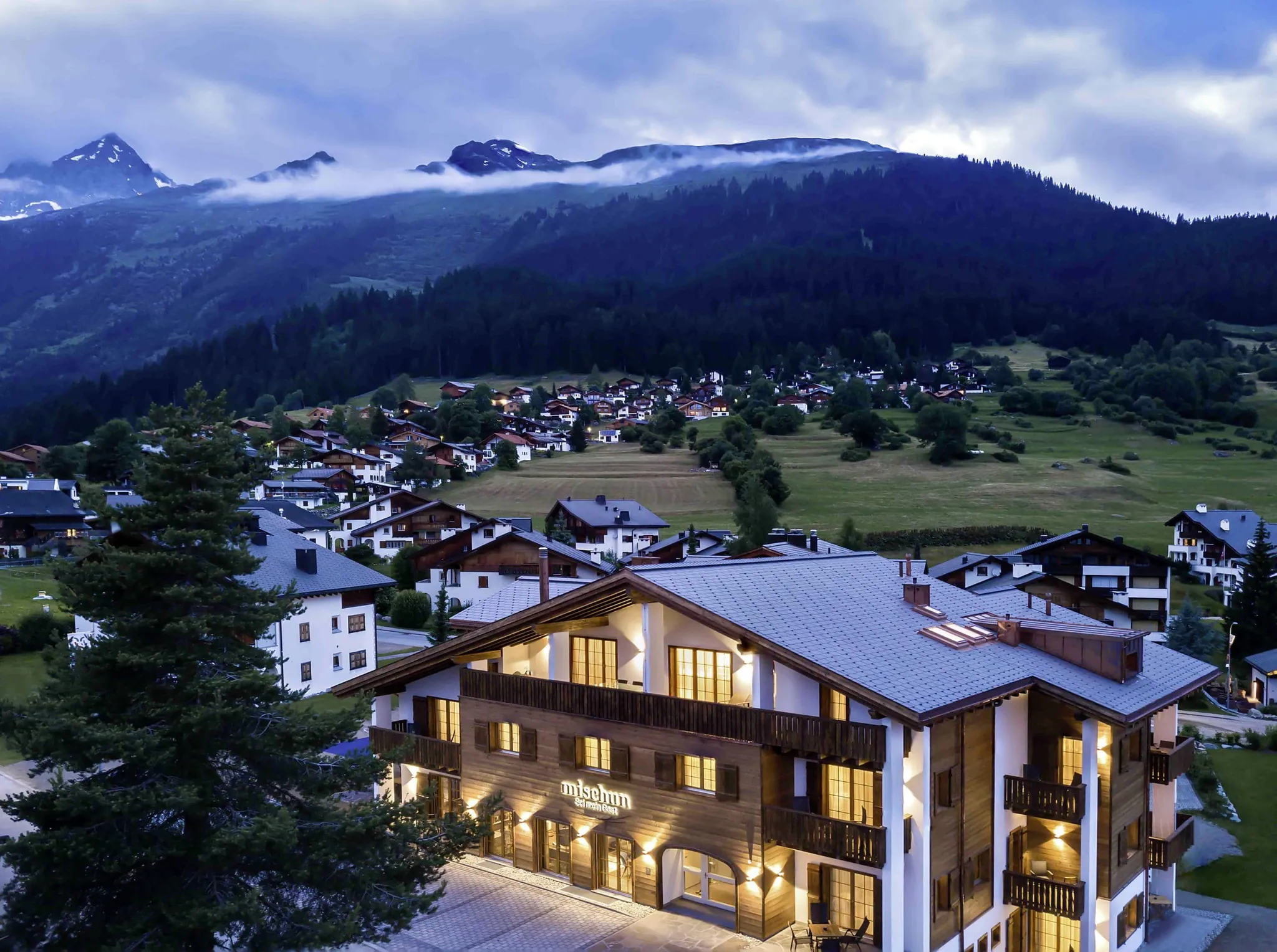 Beleuchtete Holzfassade des Hotel Mischun in einem Bergdorf, im Hintergrund bewaldete Berge unter bewölktem Himmel