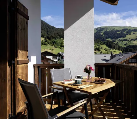Balkon im Hotel Mischun mit Holztisch, zwei Stühlen, Kaffeetassen, Muffins und Blumen, Blick auf grüne Berge und Häuser