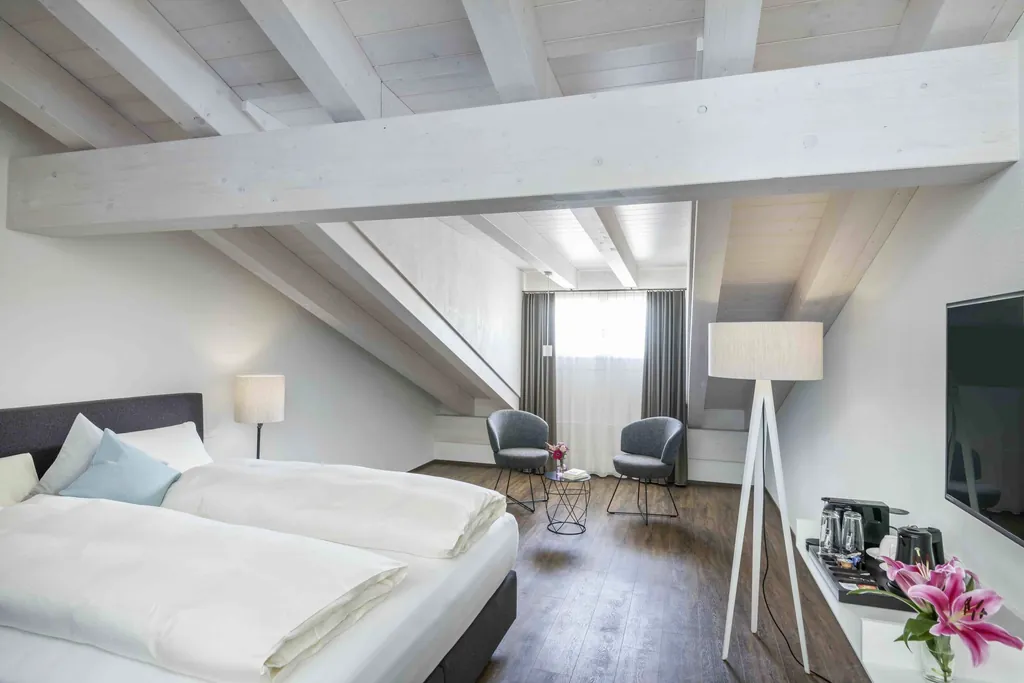 Hell eingerichtetes Schlafzimmer im Hotel Mischun mit Dachschräge, Doppelbett, zwei Sesseln, Stehlampen und Fernseher auf dunklem Holzboden