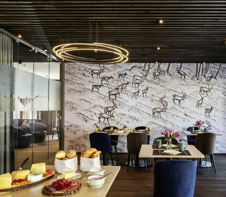 Moderner Frühstücksbereich im Hotel Mischun mit dunkler Holzdecke, runden Leuchten, blauen Stühlen und Wandbild mit Hirschherde im Schnee