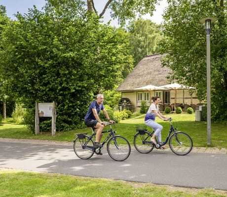 Mann und Frau fahren mit den Rädern auf einem Radweg an Mühlenhaus vorbei beim BEECH Resort Plauer See