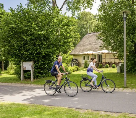 Mann und Frau fahren mit den Rädern auf einem Radweg an Mühlenhaus vorbei beim BEECH Resort Plauer See