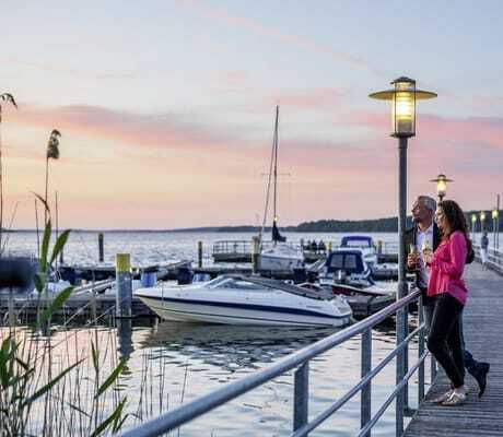 Paar steht mit Weingläsern auf dem Steg am Hafen des BEECH Resort Plauer See und schaut in den Sonnenuntergang
