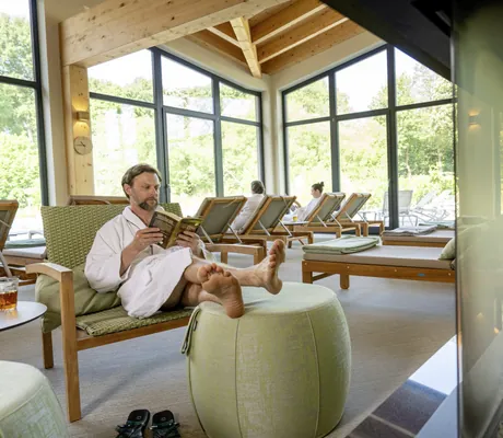 Mann liegt in weißem Bademantel auf grüner Liege am Kamin im Ruheraum des BEECH Resort Plauer See