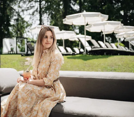 Junge Frau im Sommerkleid sitzt mit Cocktail auf einem Outdoorsofa in der Poolbar