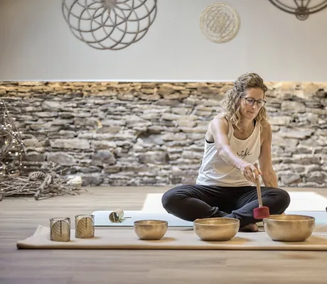 blondgelockte Frau mit Brille schlägt mit Klöppel an Klangschale im Yogaraum der Halfenstube