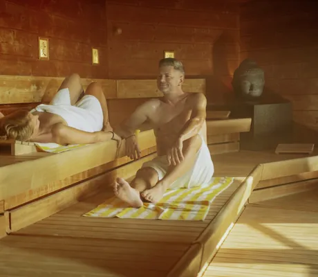 Frau liegt in der Sauna des Kurhotel an der Obermaintherme und Mann sitzt daneben auf gelb-weiß gestreiftem Badetuch