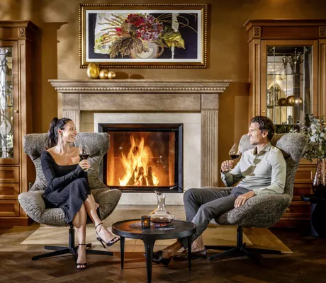 Paar sitzt in Abendkleidung mit Rotweinglas in Ohrensesseln vor dem Kamin im Mooshof