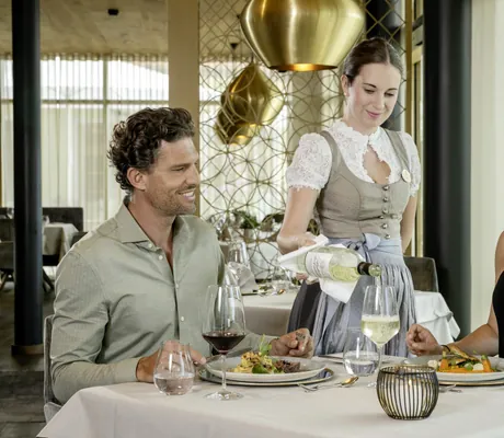 Paar sitzt im Restaurant des Mooshof beim Essen während Bedienung im Dirndl Wein einschenkt