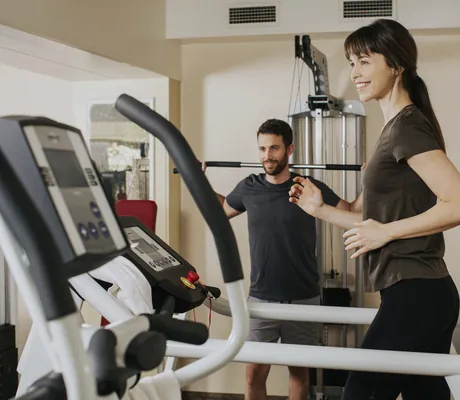dunkelhaarige Frau mit Pony läuft auf Laufband und Mann trainiert mit Hantelstange im Ortner's Resort