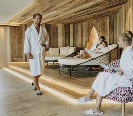 Mann in weißem Bademantel kommt zu sitzender Frau mit Dutt im Ruhebereich des Ortner's Resort