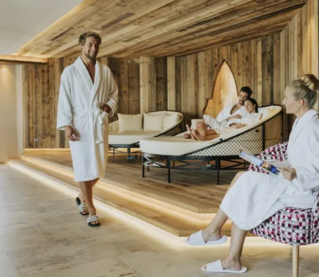 Mann in weißem Bademantel kommt zu sitzender Frau mit Dutt im Ruhebereich des Ortner's Resort
