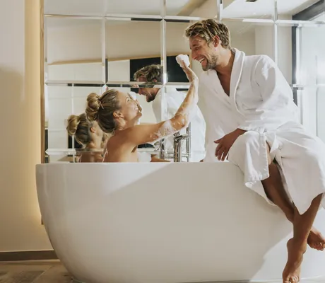 Mann in weißem Bademantel sitzt auf Badewannenrand während Frau im Wasser mit Schaum spielt im Ortner's Resort