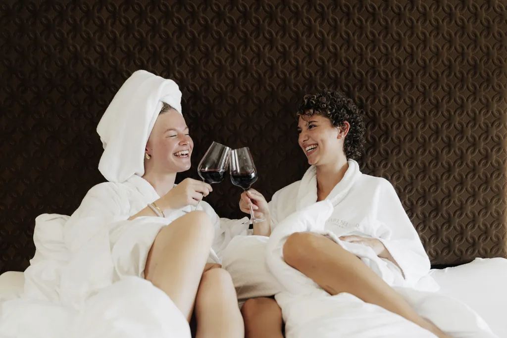 zwei Frauen in weißen Bademänteln sitzen auf dem Bett im Post Seefeld und prosten sich mit Rotwein zu