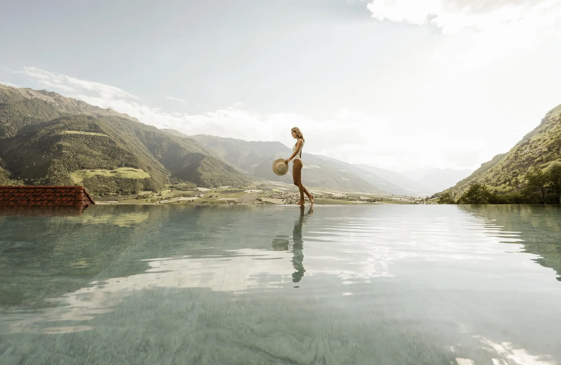 Frau in weißem Badeanzug steht mit Hut in der Hand am Infinitypool des Preidlhof