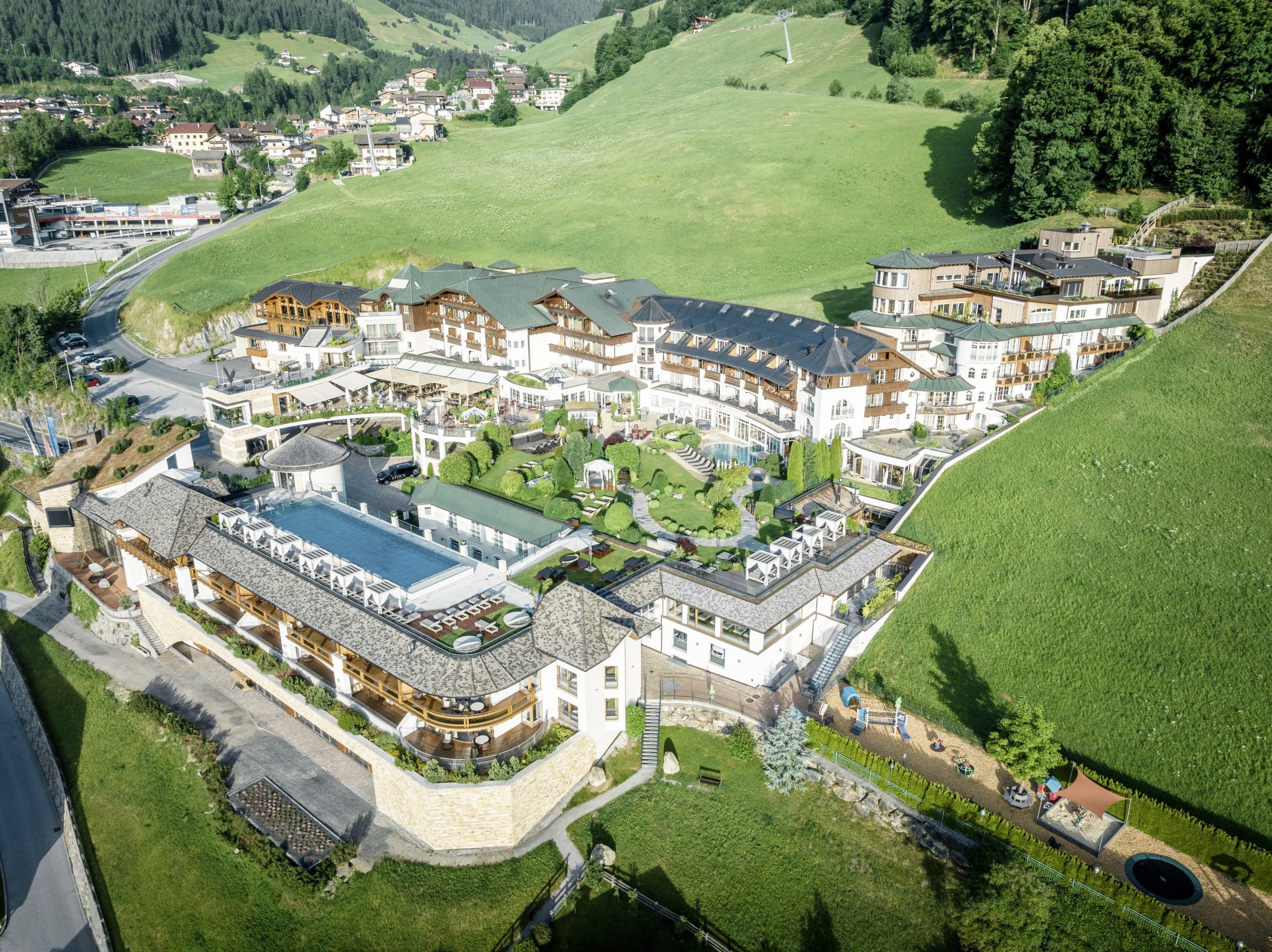 Luftaufnahme der Hotelanlage des STOCK resort mit mehreren Gebäuden, Außenpool und umliegenden grünen Wiesen und Wäldern