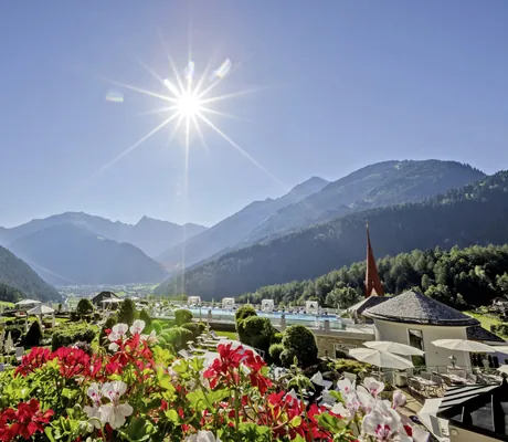 Blühende rote und weiße Blumen, im Hintergrund Gartenanlage des STOCK resort mit einigen Liegestühlen, Bergen und strahlende Sonne am blauen Himmel