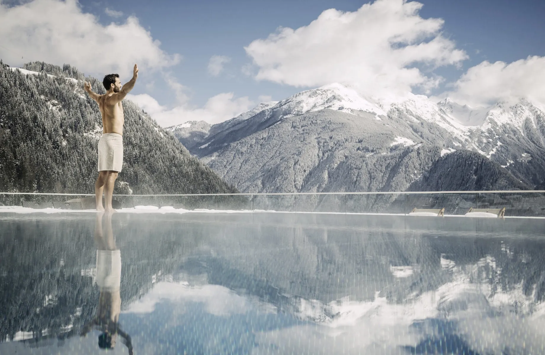 Mann mit Badehose steht am Rand des Outdoorpools des STOCK resort vor schneebedeckten Bergen und breitet die Arme aus