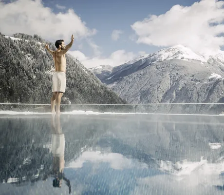 Mann mit Badehose steht am Rand des Outdoorpools des STOCK resort vor schneebedeckten Bergen und breitet die Arme aus