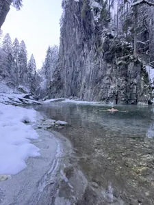Mann mit ausgebreiteten Armen in klarem Flusswasser beim Eisbaden, umgeben von schneebedeckten Felsen und steilen Felswänden in der Nähe des STOCK resort