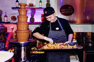 Koch des STOCK resort in schwarzer Kleidung und Schürze bereitet Obststücke vor, daneben große Schokoladenfontäne, im Hintergrund Schild mit Text 'Aber bitte mit Sahne'