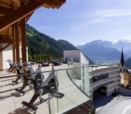 Sonnige Terrasse des STOCK resort mit mehreren Fitnessrädern, Blick auf Berglandschaft und Kirchturm bei klarem Himmel