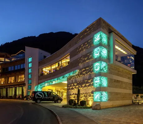 Beleuchtete Hotelfassade des STOCK resort mit traditionellem und modernem Teil vor Bergkulisse bei Abenddämmerung, Fahrzeug mit Hotel-Logo vor Einfahrt