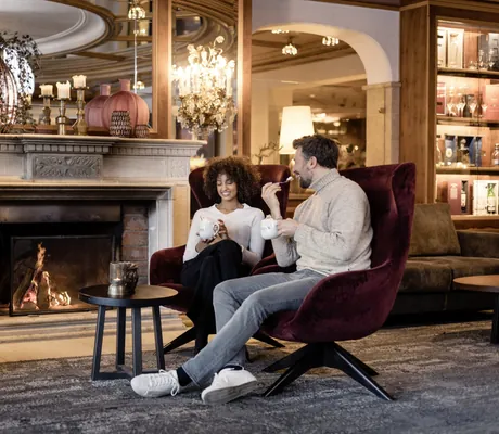 Zwei Personen sitzen in dunkelroten Samtsesseln vor einem Kamin mit brennendem Feuer im STOCK resort, beide halten eine Tasse in der Hand