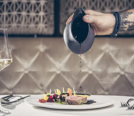 Hand mit Armband gießt dunkle Sauce aus kleinem Krug auf Teller mit Fleisch und Gemüse im Restaurant des STOCK resort