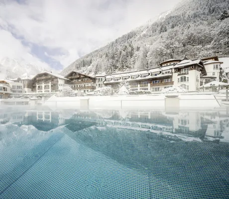 Outdoorpool des STOCK resort vor schneebedeckten Bergen und Hotelfassade im Hintergrund