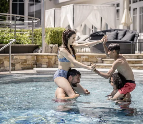 Zwei Kinder im Outdoorpool des STOCK resort, die auf den Schultern von Erwachsenen sitzen und sich an den Händen halten