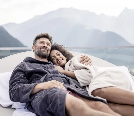 Paar liegt in Bademänteln auf einer Outdoor-Liege des STOCK resort mit Bergpanorama im Hintergrund