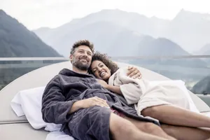 Paar liegt in Bademänteln auf einer Outdoor-Liege des STOCK resort mit Bergpanorama im Hintergrund