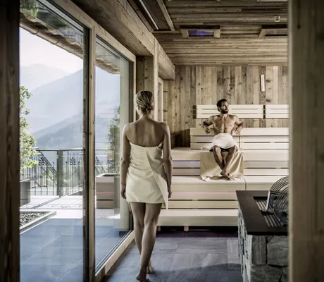 Frau mit weißem Handtuch steht in einer Sauna im STOCK resort, Mann sitzt auf Holzbank, große Fenster mit Bergblick