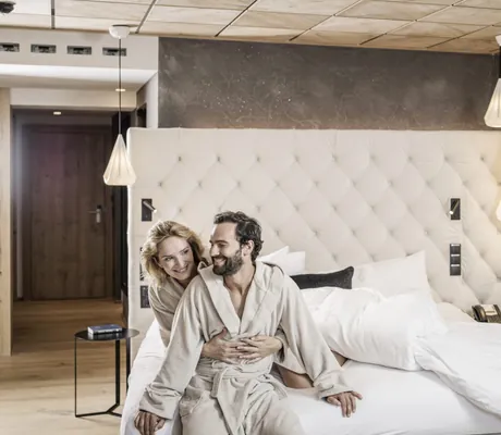 Paar in beigefarbenen Bademänteln sitzt auf Bett im STOCK resort mit gepolstertem Kopfteil in modernem Schlafzimmer mit Holzdecke und hängenden Lampen