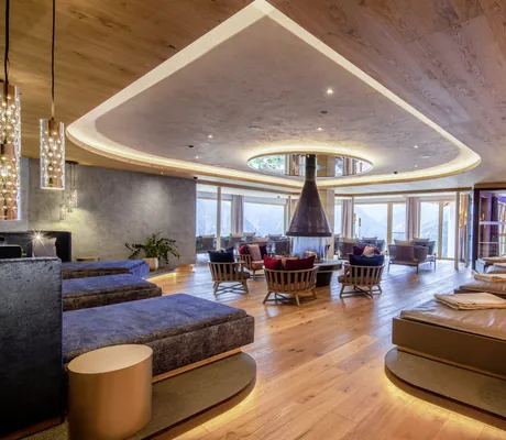 Großer, moderner Panoramaruheraum im STOCK resort mit Holzdecke und -boden, zentralem Kamin, Sessel und Liegen, großen Fenstern mit Bergblick