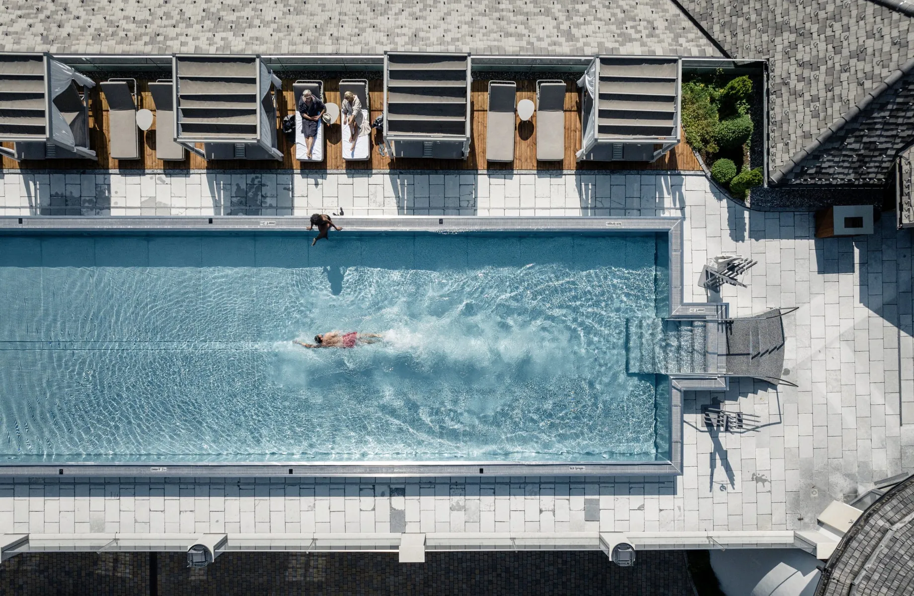 Luftaufnahme des Outdoorpools im STOCK resort mit schwimmender Person, Liegen und Himmelbetten am Beckenrand
