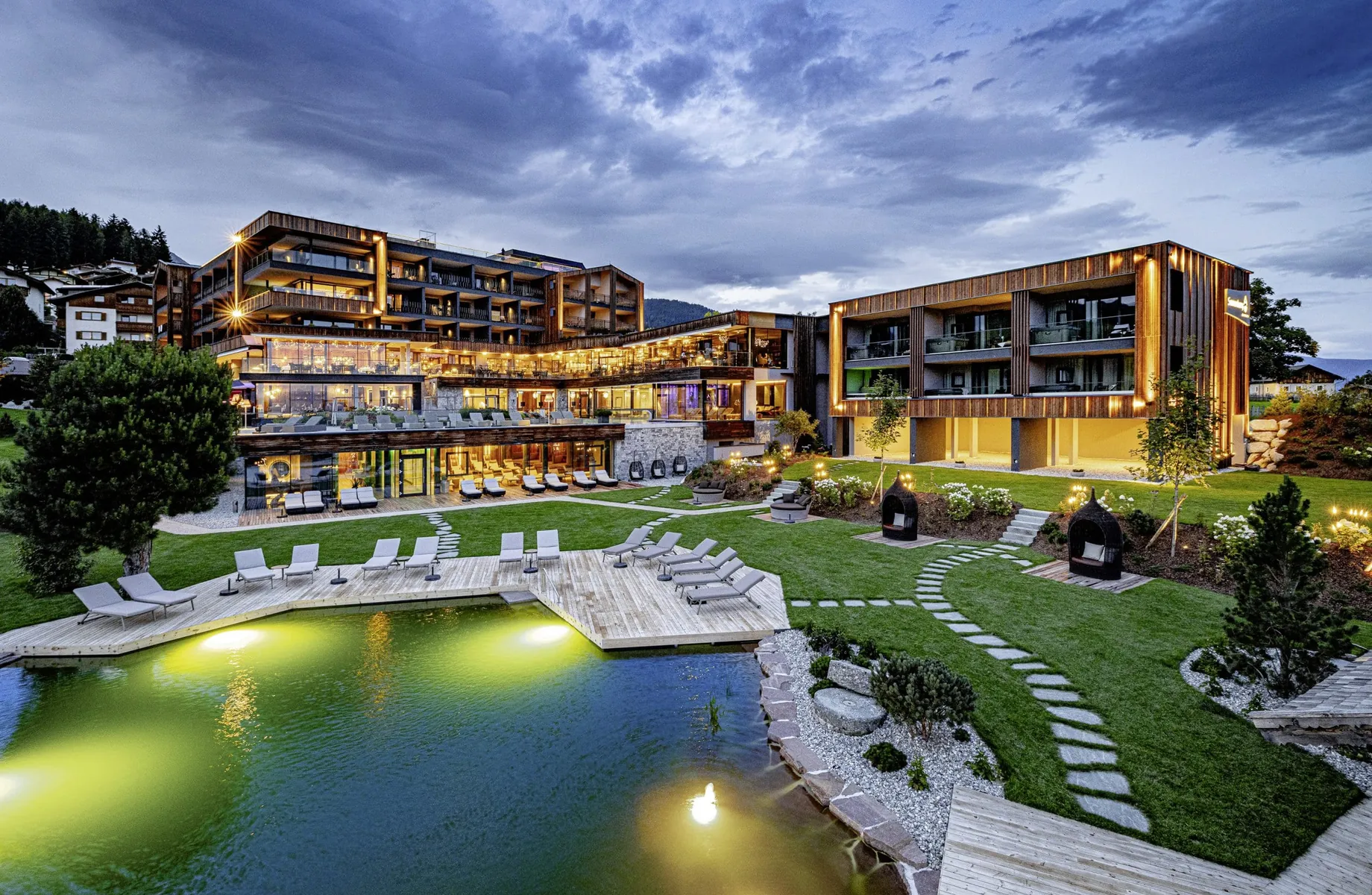 Gesamtansicht des Sonnenberg Panorama Dolomites Wellbeing vom Naturbadeteich aus in er Abenddämmerung