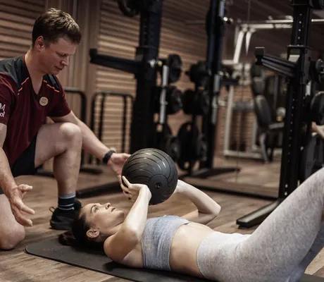 Fitnesstrainer des Tuxerhof hockt hinter einer Frau auf einer Gymnastikmatte um einen Ball zu fangen