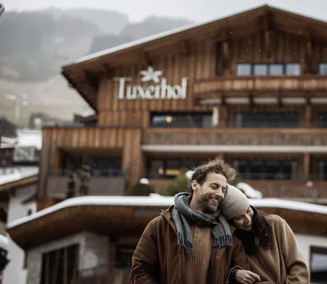 Paar steht eingehakt lachend vor der Fassade des Tuxerhof