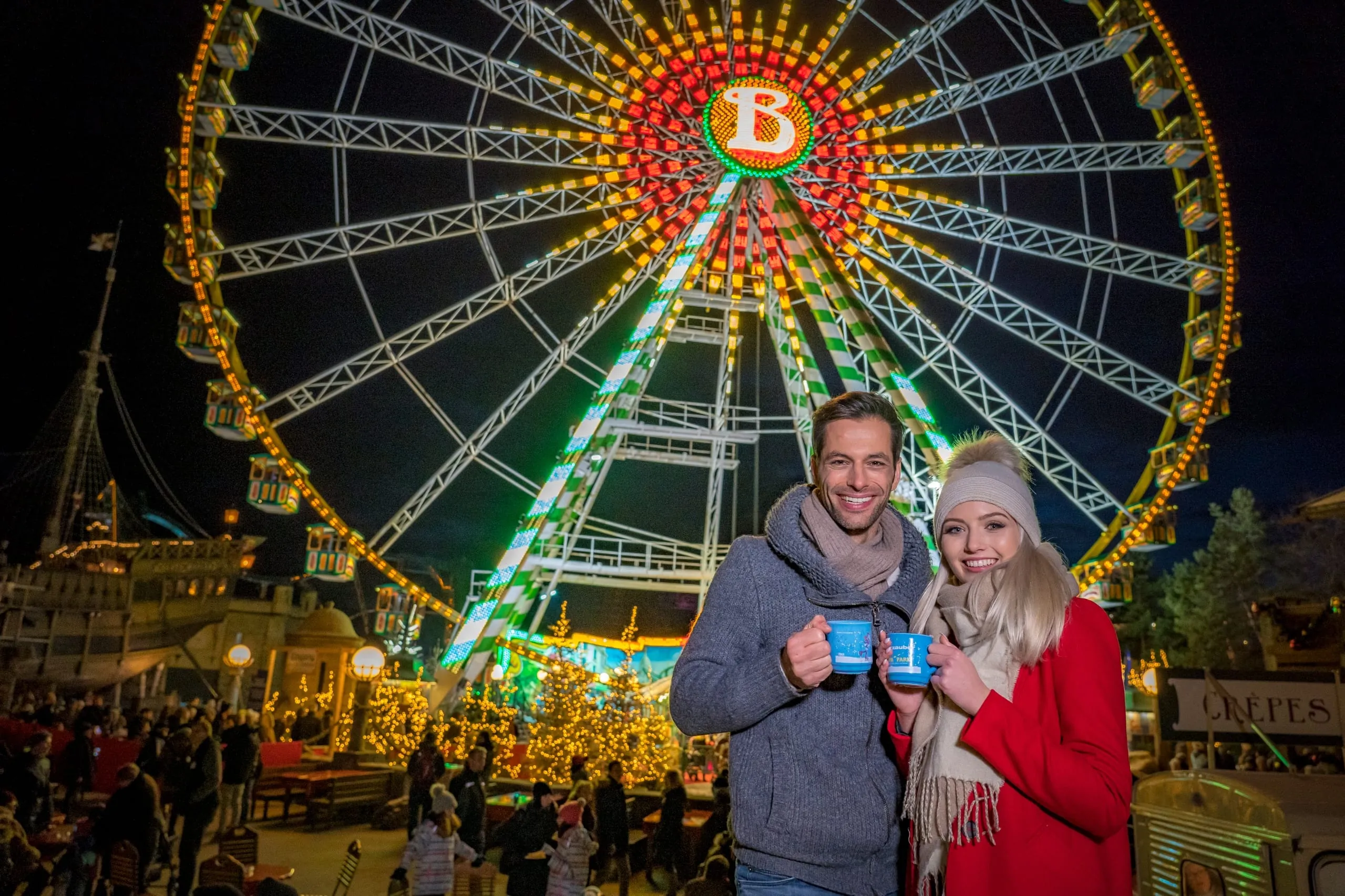 Winterlich bekleidetes Pärchen steht mit Glühweintassen in der Hand vor dem beleuchteten Riesenrad im Europa-Park in Rust